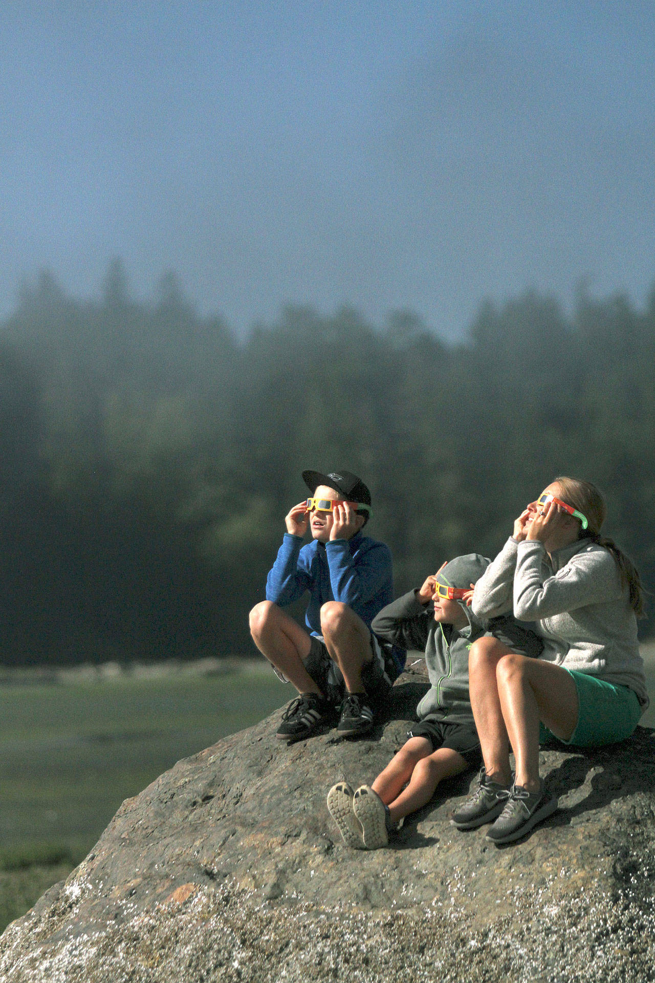 Brian Kelly | Bainbridge Island Review                                Tamra Hauge (far right) watches the eclipse with her sons Magnus and Alden from the perch of a big rock at Manitou Beach.