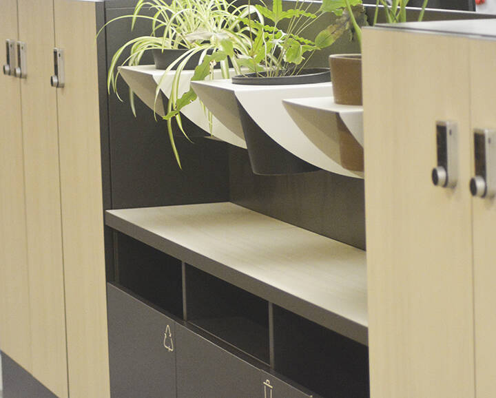 This piece of furniture includes lockers left and right with recycling bins in the middle and plants in the middle and is located near workers for convenience.