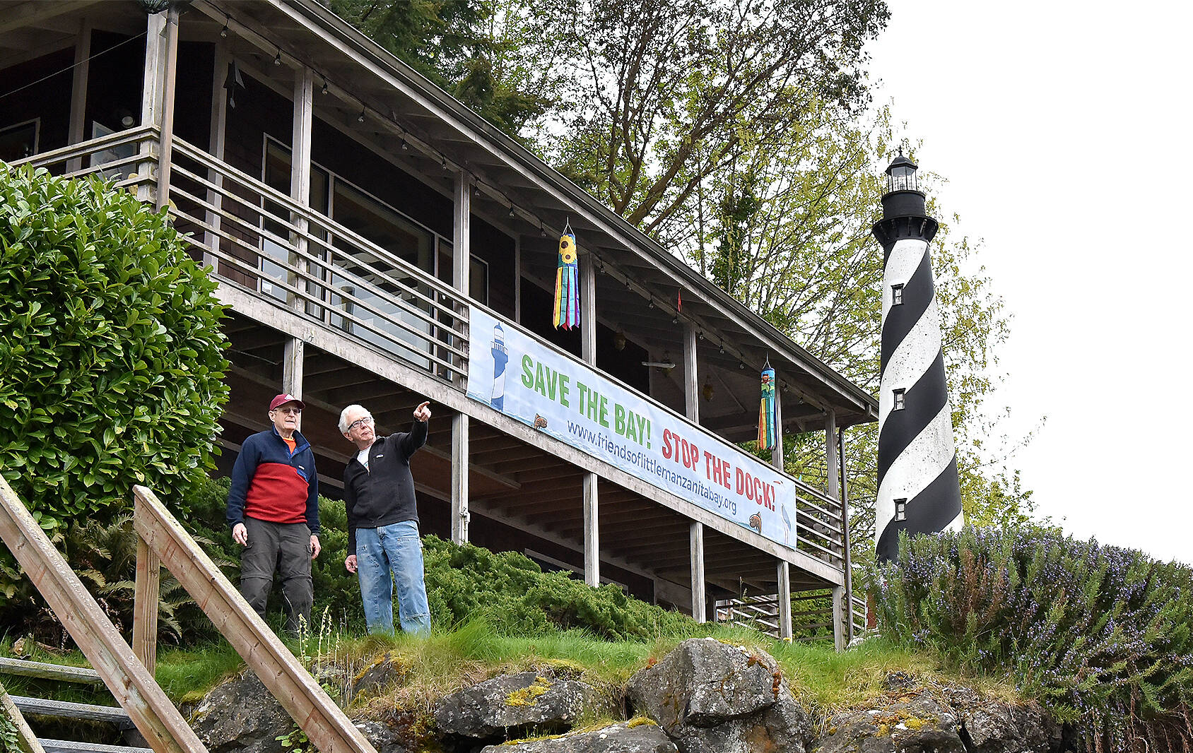 Dr. Fred Grimm and Jim Coon, president of the board of Friends of Little Manzanita Bay, stand beneath a sign they put up to protest a proposal submitted by two neighbors who want to replace an 84-foot wooden dock with a 240-foot steel pier, ramp and dock including two boat lifts. Grimm, who has lived on the point with his wife, Willie since the 1970s, decided to display their protest by hanging the 25-foot-long banner in front of their house with the words, “SAVE THE BAY! STOP THE DOCK!”  Little Manzanita Bay is designated Critical Habitat, under the Endangered Species Act, for Puget Sound Chinook salmon, Puget Sound Rockfish, and Southern Resident Killer Whale. The Bay is also considered Essential Fish Habitat for Coho, Chinook, and Puget Sound pink salmon.Nancy Treder/Bainbridge Island Review