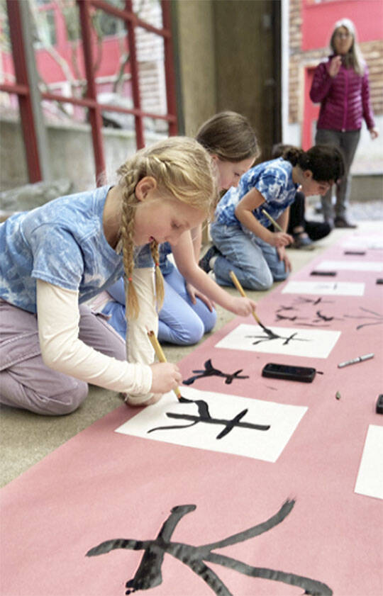 Second-graders practice brush strokes for the Japanese symbol of ‘tree.’ The children are wearing shirts they made with indigo dye in the style of shibori, which means to wring, squeeze and press.
