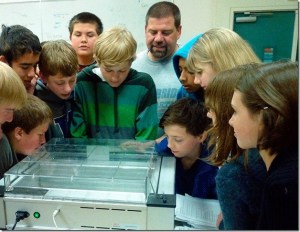 Technology students in Sean Eaton’s class at Woodward Middle School gather around their teacher to look at the classroom’s new 2BOT-3D printer “etching” out a shape that they sent to the device via their computer.