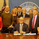 Gov. Jay Inslee, seated, signs the first bill of the 2020 legislative session into law. On the right stands the bills primary sponsor, Sen. Jamie Pedersen, D-Seattle, who is wearing a red tie. (Cameron Sheppard | WNPA News Service)