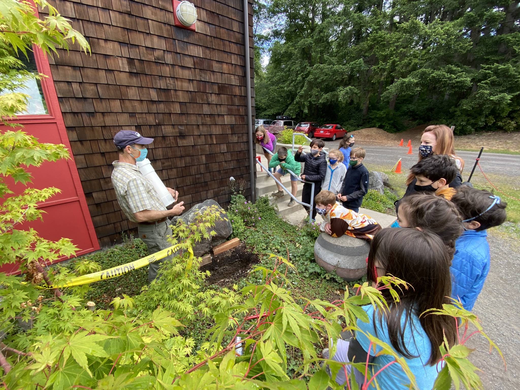 Joan Henderson courtesy photo
Island School students ask questions of teacher Mike Derzon, who holds a time capsule near the deep hole in the garden where they buried it, to be opened by students in 2046. To commemorate a COVID-19 school experience, children included: hand warmers for the outdoor classrooms, a school newspaper written by 4th- and 5th- graders, dolphin and blue marlin art for two pod mascots, class photos with masked faces, and a hummingbird nest to signify the 2nd and 3rd grade outdoor bird study. Kindergartners crafted a coloring book of their memories. Others popped in hand sanitizer, gloves and a facemask to tell future students of the safety measures they took during the pandemic.