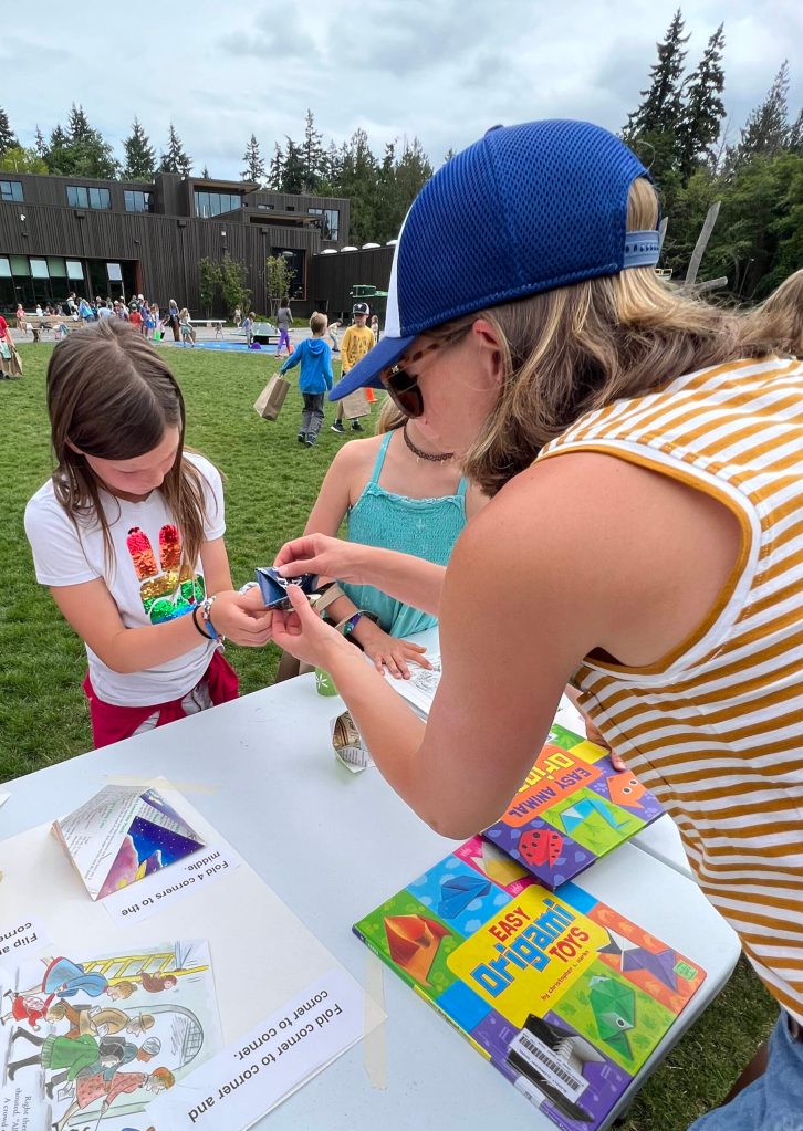 A parent volunteer helps a student make a folded fortune teller paper toy.