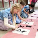 Second-graders practice brush strokes for the Japanese symbol of tree. The children are wearing shirts they made with indigo dye in the style of shibori, which means to wring, squeeze and press.